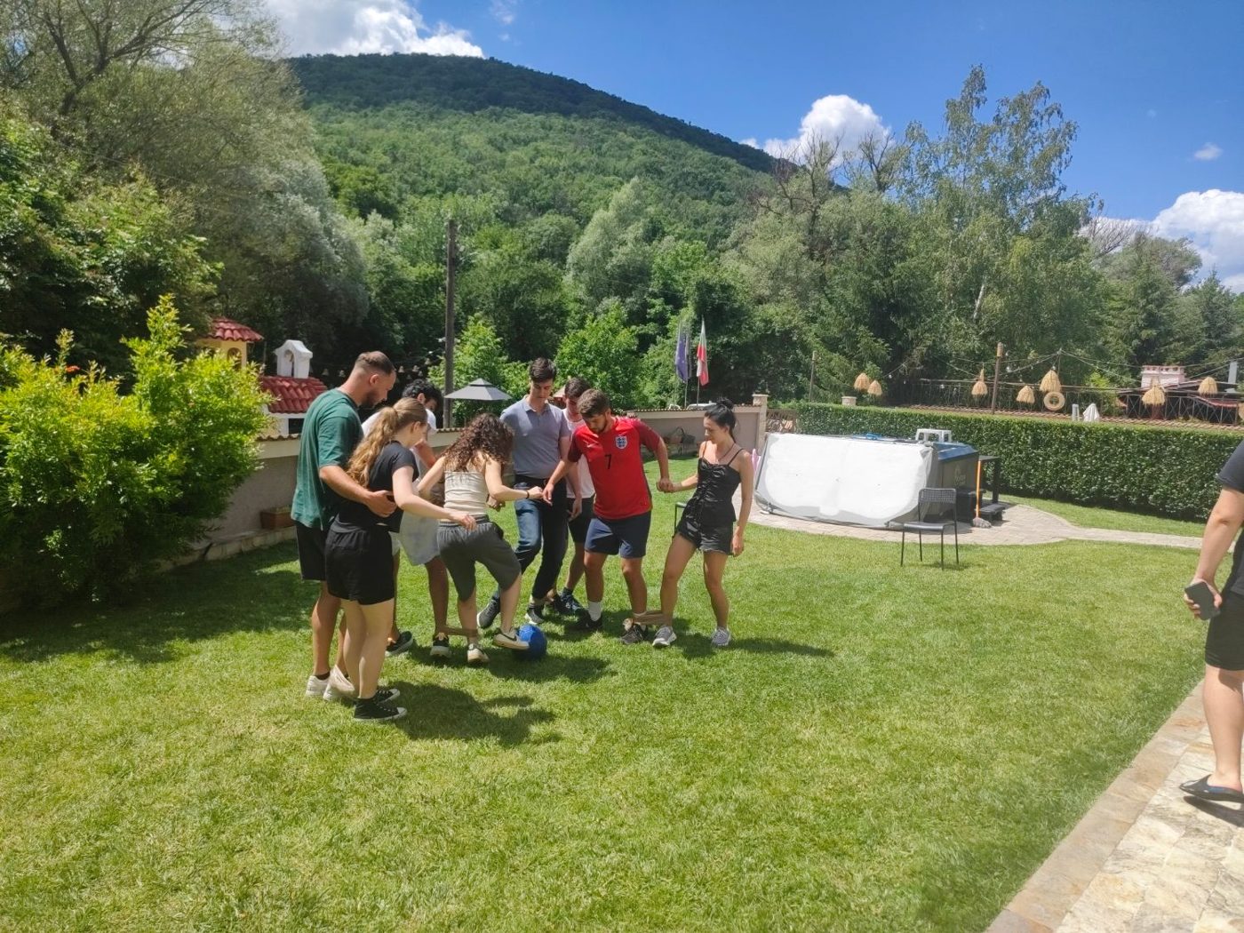 Participants with their ankles tied together huddle around a football in the garden, a green mountain behind them